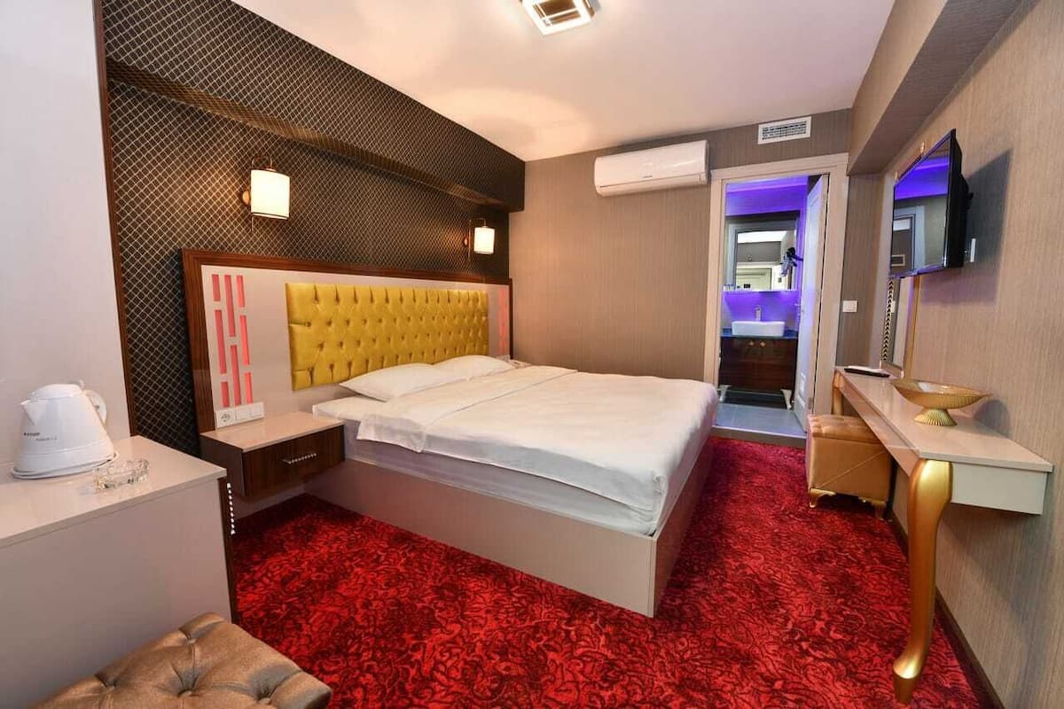 Habitación de hotel con cabecera amarilla con botonadura, alfombra roja estampada, mesita de noche con hervidor blanco, un taburete pequeño y un tocador con espejo y un bol.