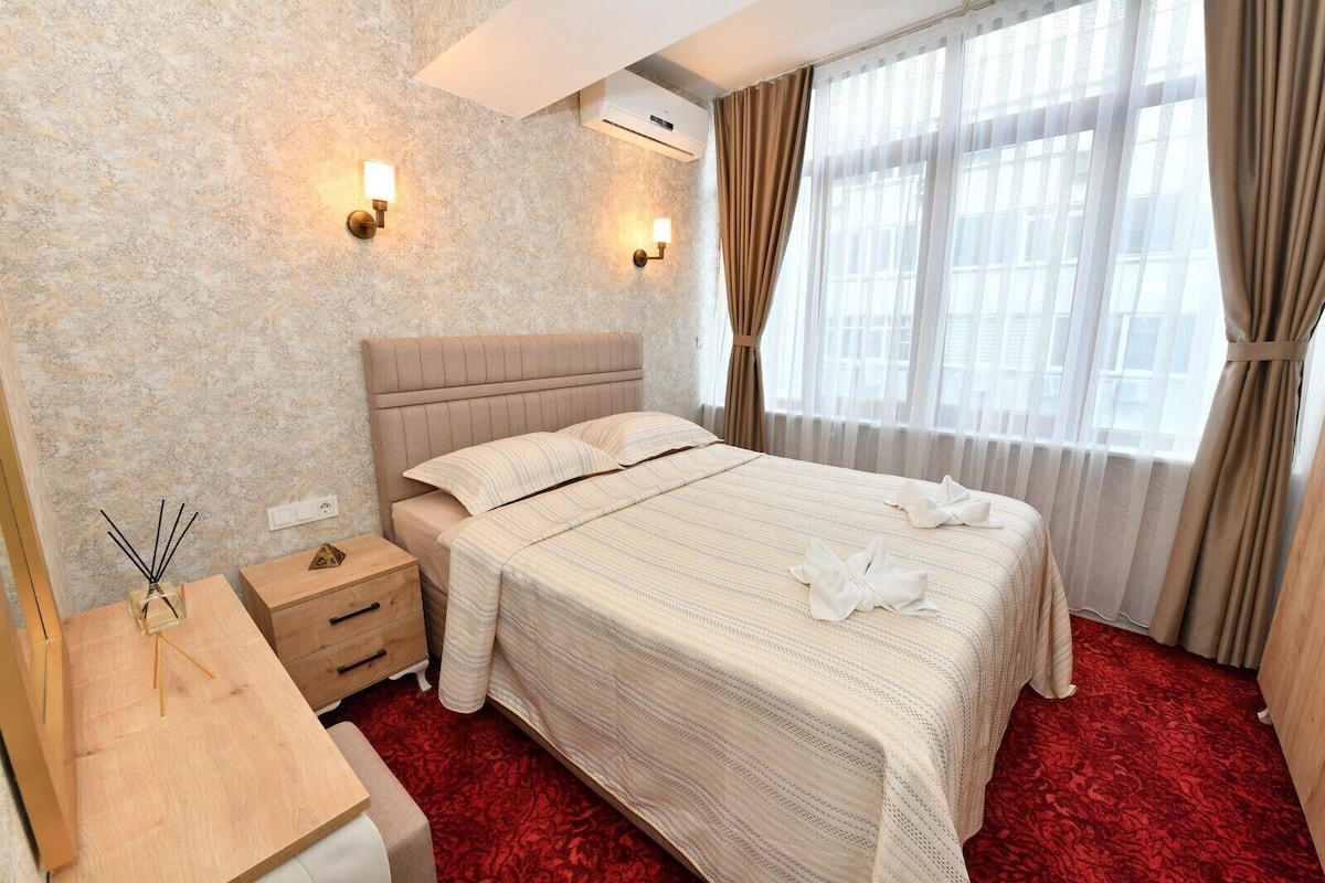 Habitación de hotel con cama, mesitas de noche, aire acondicionado instalado en la pared y una ventana con cortinas.