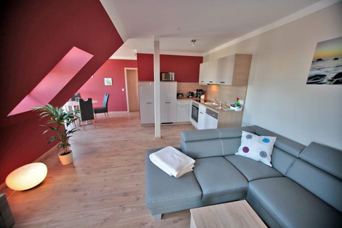 Un appartement moderne avec un canapé gris, un coin repas, une cuisine et un mur d’accent rouge.