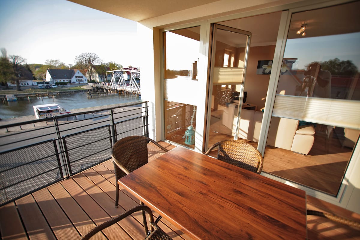 Un balcon avec une table en bois et des chaises en osier, donnant sur une étendue d’eau où l’on peut voir des bateaux et un quai.