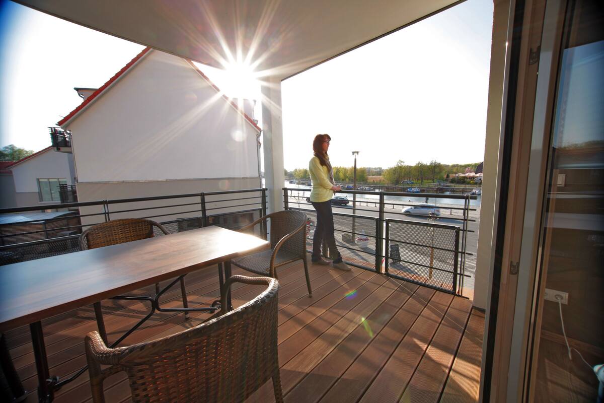 Un balcon avec une table et des chaises, donnant sur un parking et une étendue d’eau.