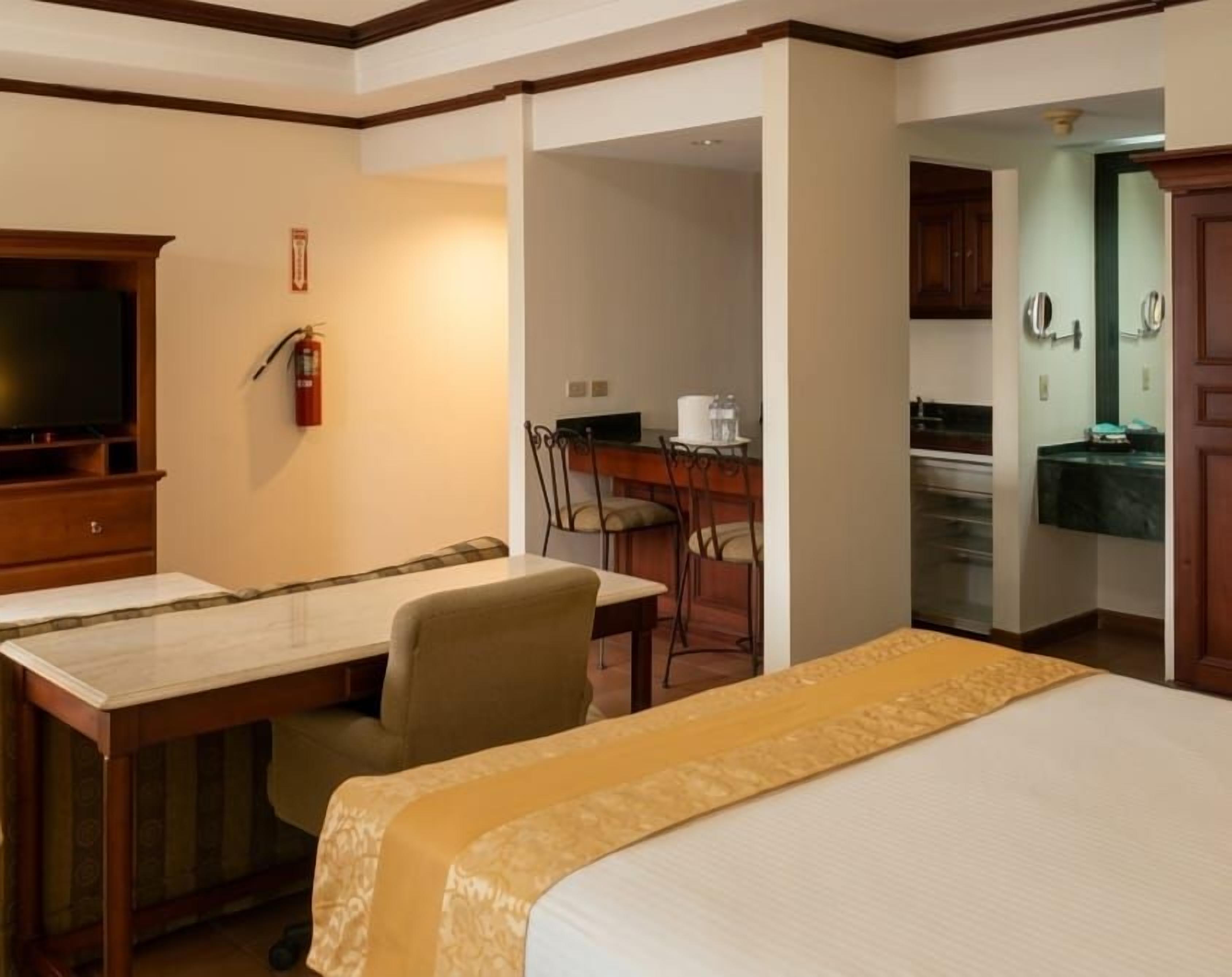 A hotel room with a bed, a desk, a chair, a television, and a kitchenette area.