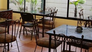 Un restaurante con mesas y sillas de vidrio, piso de baldosas y plantas en macetas junto a las ventanas.