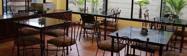 Un restaurante con mesas y sillas de vidrio, piso de baldosas y plantas en macetas junto a las ventanas.