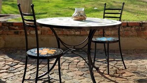 Una mesa redonda exterior con cubierta de mosaico, dos sillas con asientos de mosaico y un tarro de vidrio sobre la mesa.