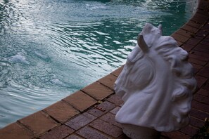 Une statue blanche se dresse près d’une piscine bordée de briques, dont l’eau est légèrement agitée.