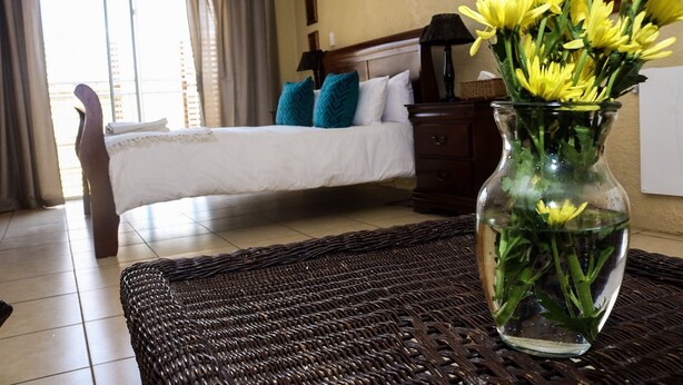 Habitación de hotel con una cama, una mesita de noche, una lámpara y un florero con flores sobre un tapete tejido.