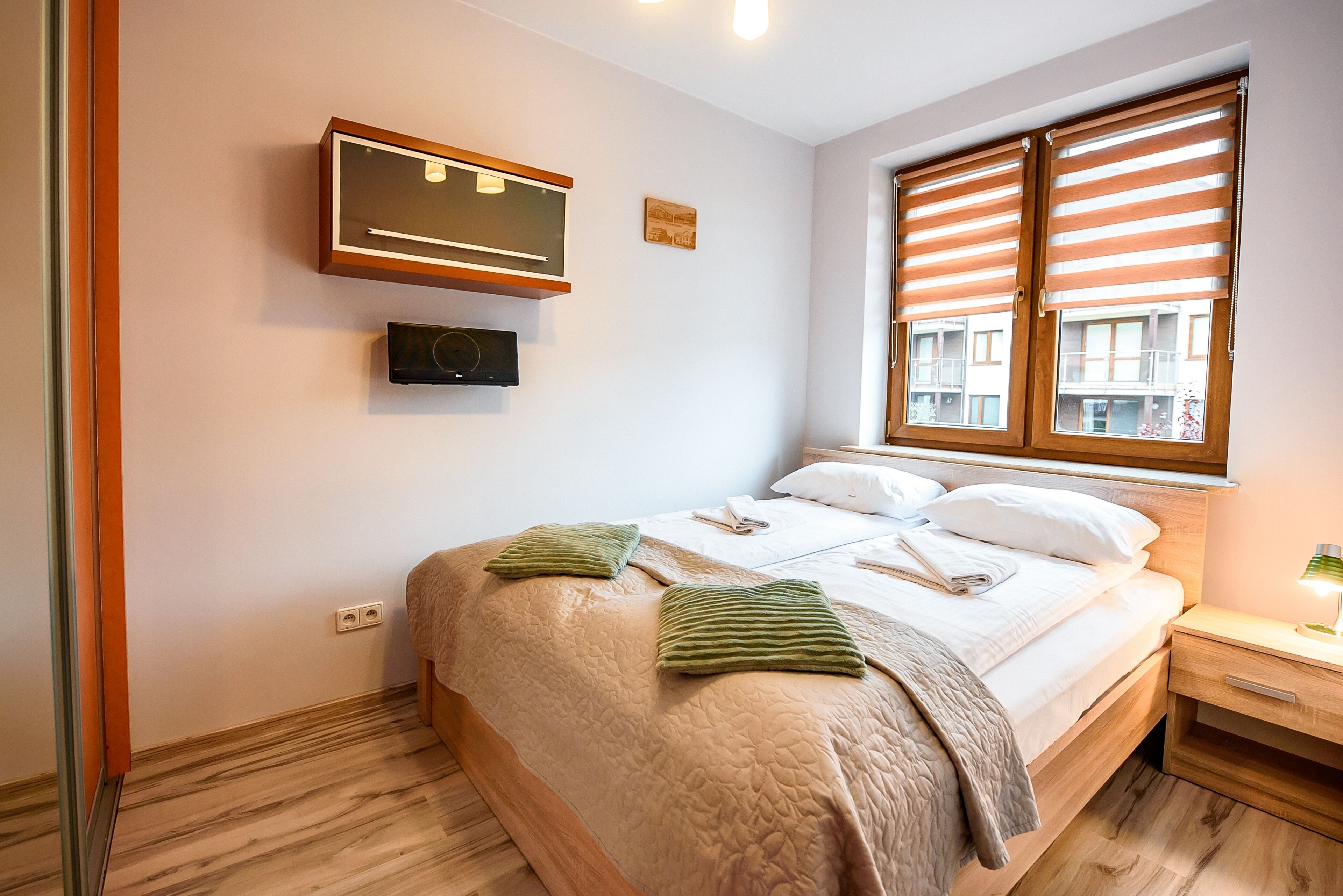 A neatly made bed with a quilt, pillows, and a blanket, a bedside table with a lamp, and a window with blinds.