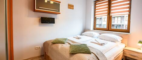 A neatly made bed with a quilt, pillows, and a blanket, a bedside table with a lamp, and a window with blinds.