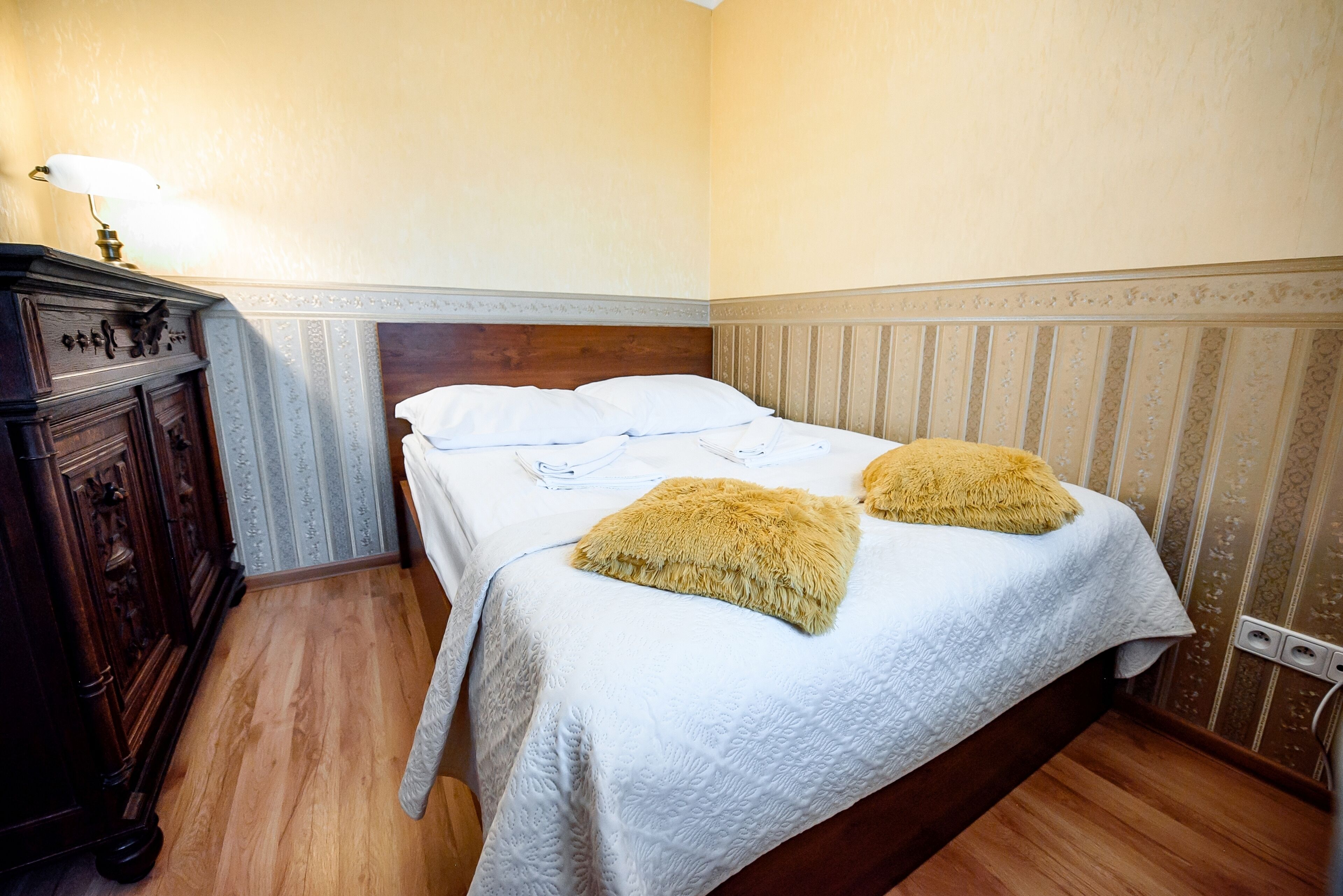 A hotel room with a wooden floor, a bed with white bedding and two fluffy pillows, a nightstand with a lamp, and a wooden dresser.
