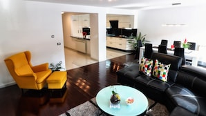 Una sala moderna con un sillón amarillo, un sofá negro con cojines coloridos y una mesa redonda de centro con un florero y un cuenco.