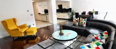 A modern living room with a sofa, armchair, and coffee table. A kitchen is visible in the background.