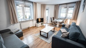 A modern living room with a sofa, a coffee table, and a TV. There is a dining area with chairs and a table. The room has large windows with curtains. - Apartamenty Sun & Snow Nad Potokiem (Krynica-Zdroj)