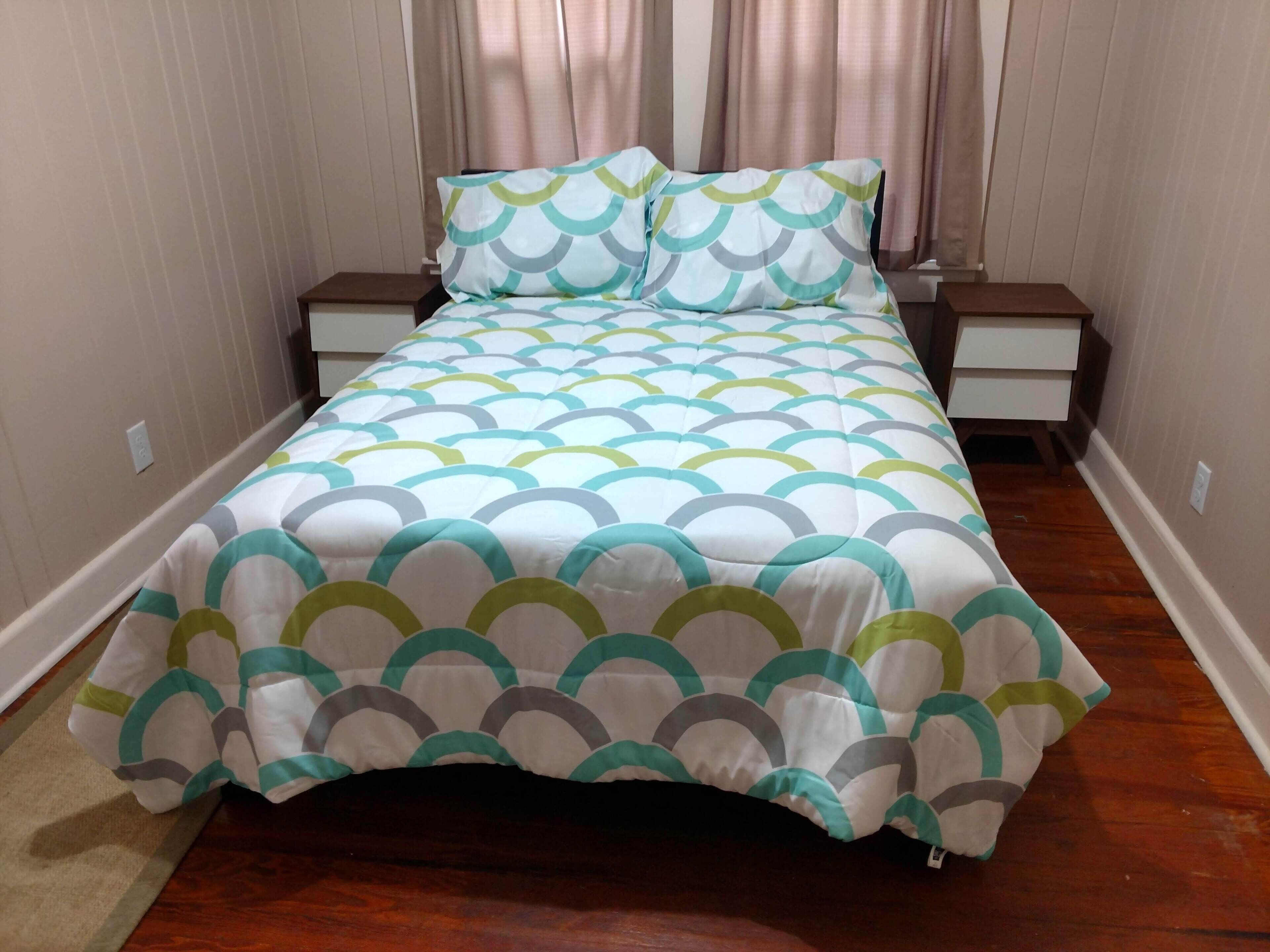 A neatly made bed with a geometric patterned bedspread and matching pillows, flanked by bedside tables with lamps, in a room with wooden flooring and light-colored walls.