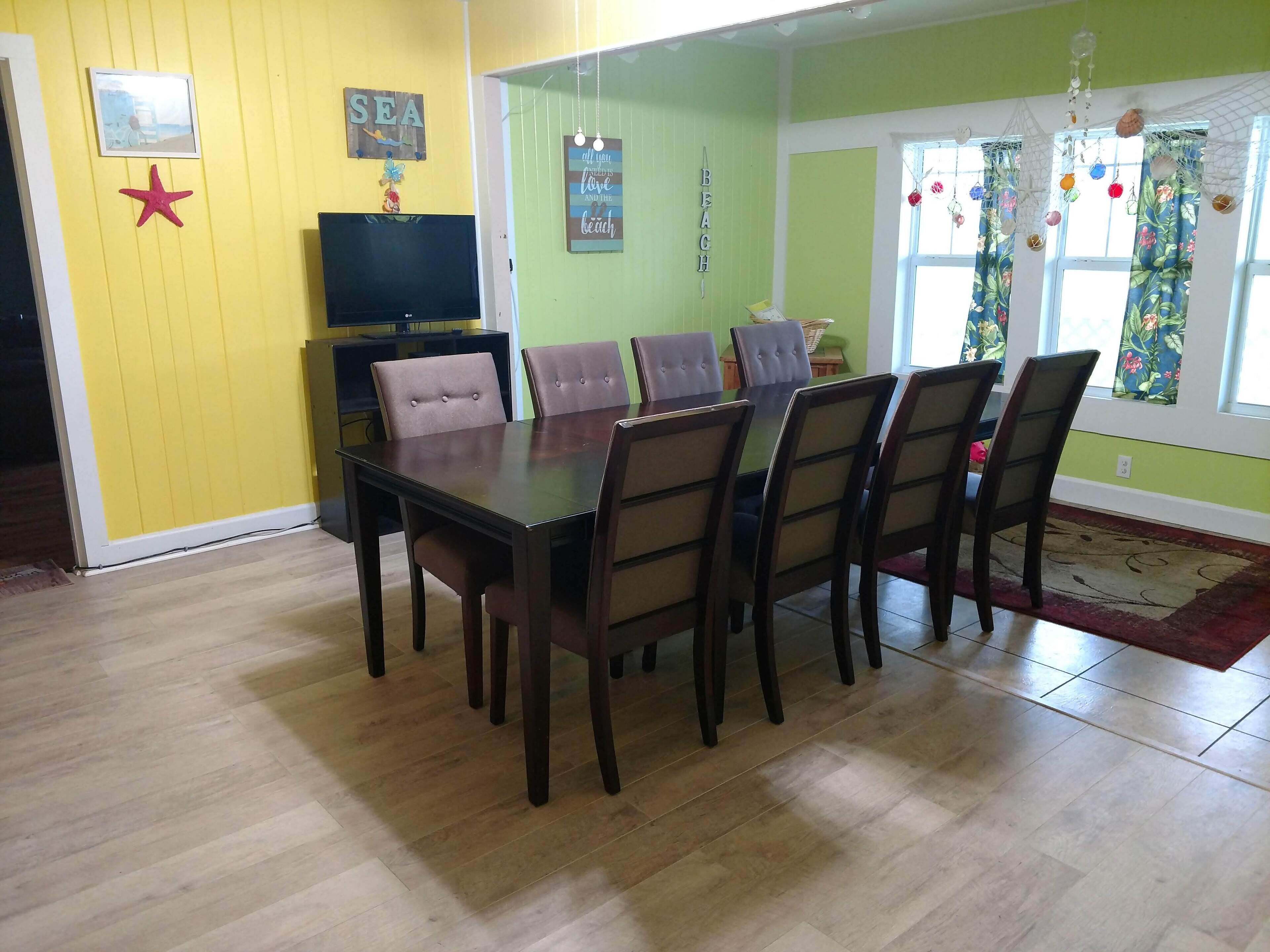 A dining area with a wooden table and chairs, a television on a stand, and a wall with a 'SEA' sign and a 'BEACH' sign.