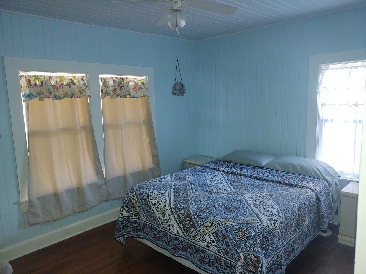 Un dormitorio con cama, dos ventanas con cortinas, un ventilador de techo y una mesita de noche pequeña.