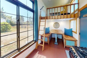 A cozy cabin with a loft bed, a dining table with blue cushions, and a view of the outdoors through large windows.