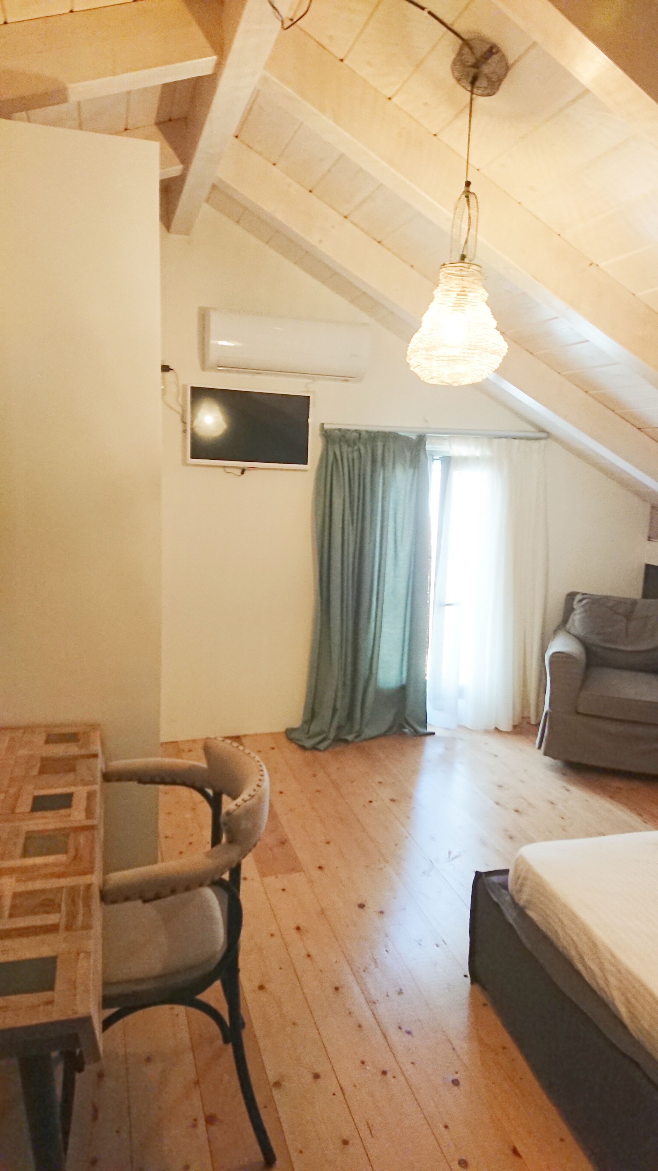 Una habitación con piso de madera, una cama, una mesa con sillas, un televisor y una ventana con cortinas.