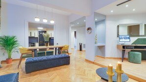 A modern living room with a sofa, a dining area with chairs, a kitchen in the background, and a green ottoman. - AthiNN Residence (Athens)