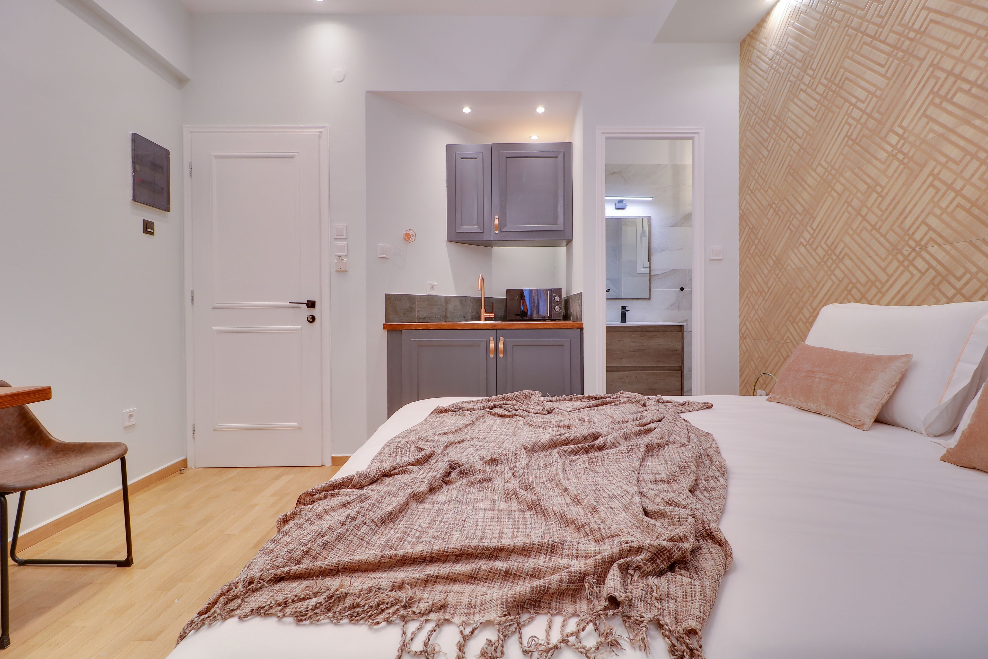 A bedroom with a bed, a chair, a kitchenette, and a bathroom visible through an open door.