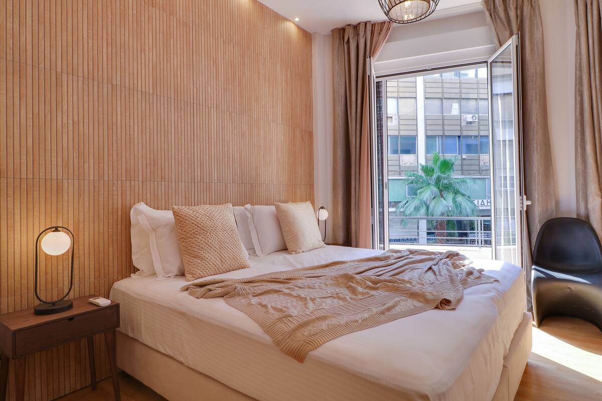 A modern hotel room with a large bed, a bedside table with a lamp, a view of the outdoors through a sliding door, and a chair by the window.