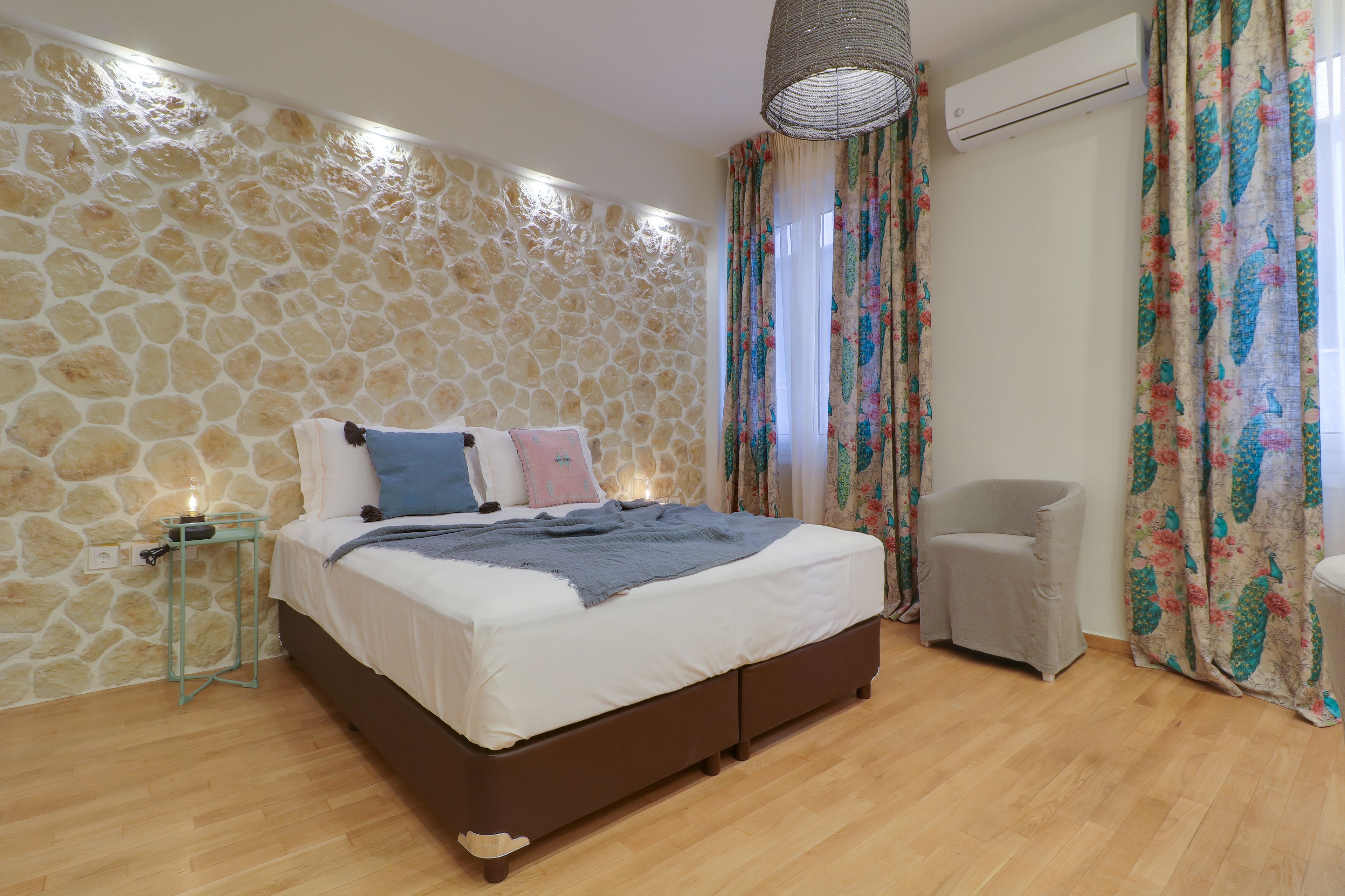 A bedroom with a stone wall, a bed with a white and blue bedspread, a grey chair, and a hanging light fixture.