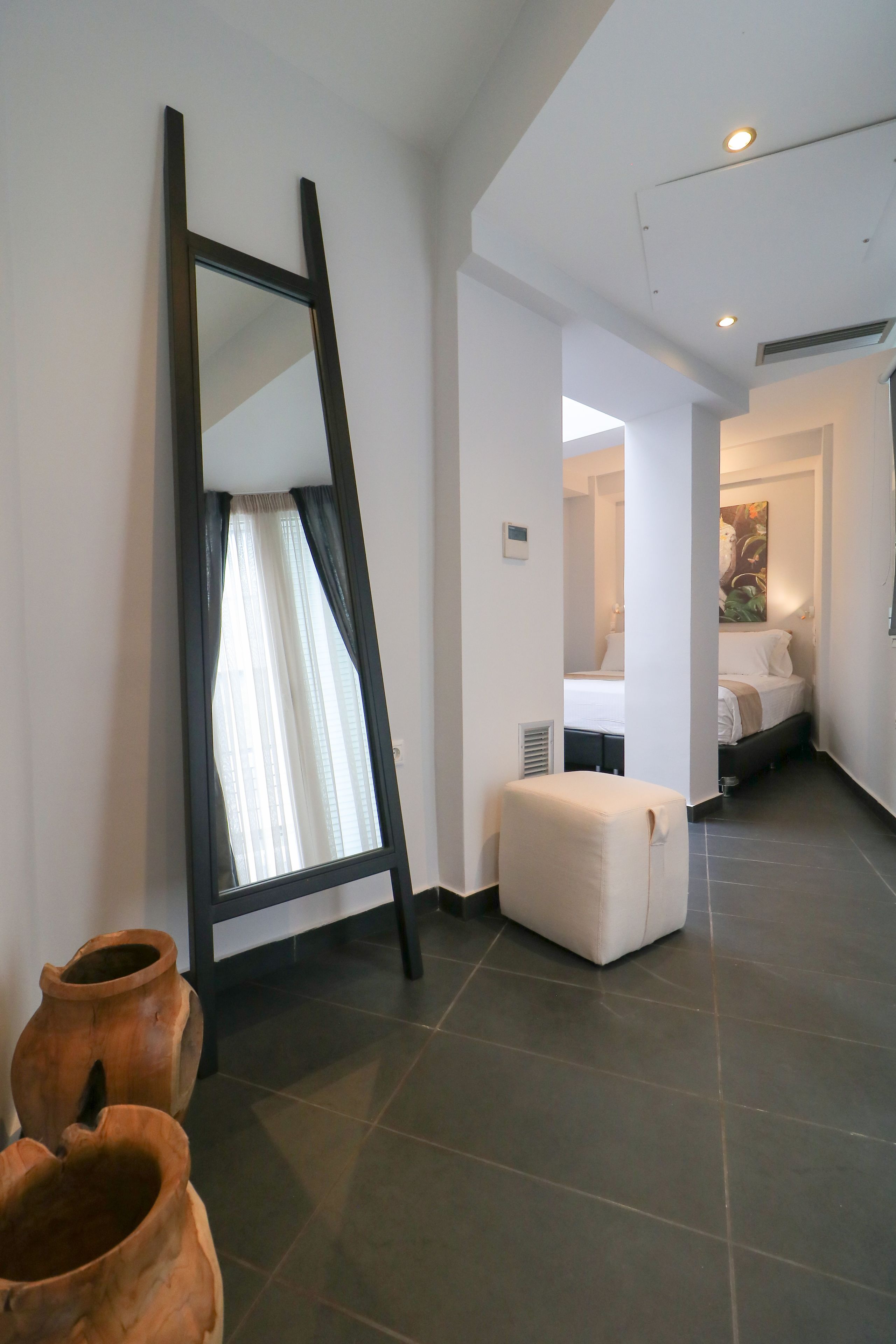 A modern hotel room with a large mirror, a white ottoman, and a bed in the background.