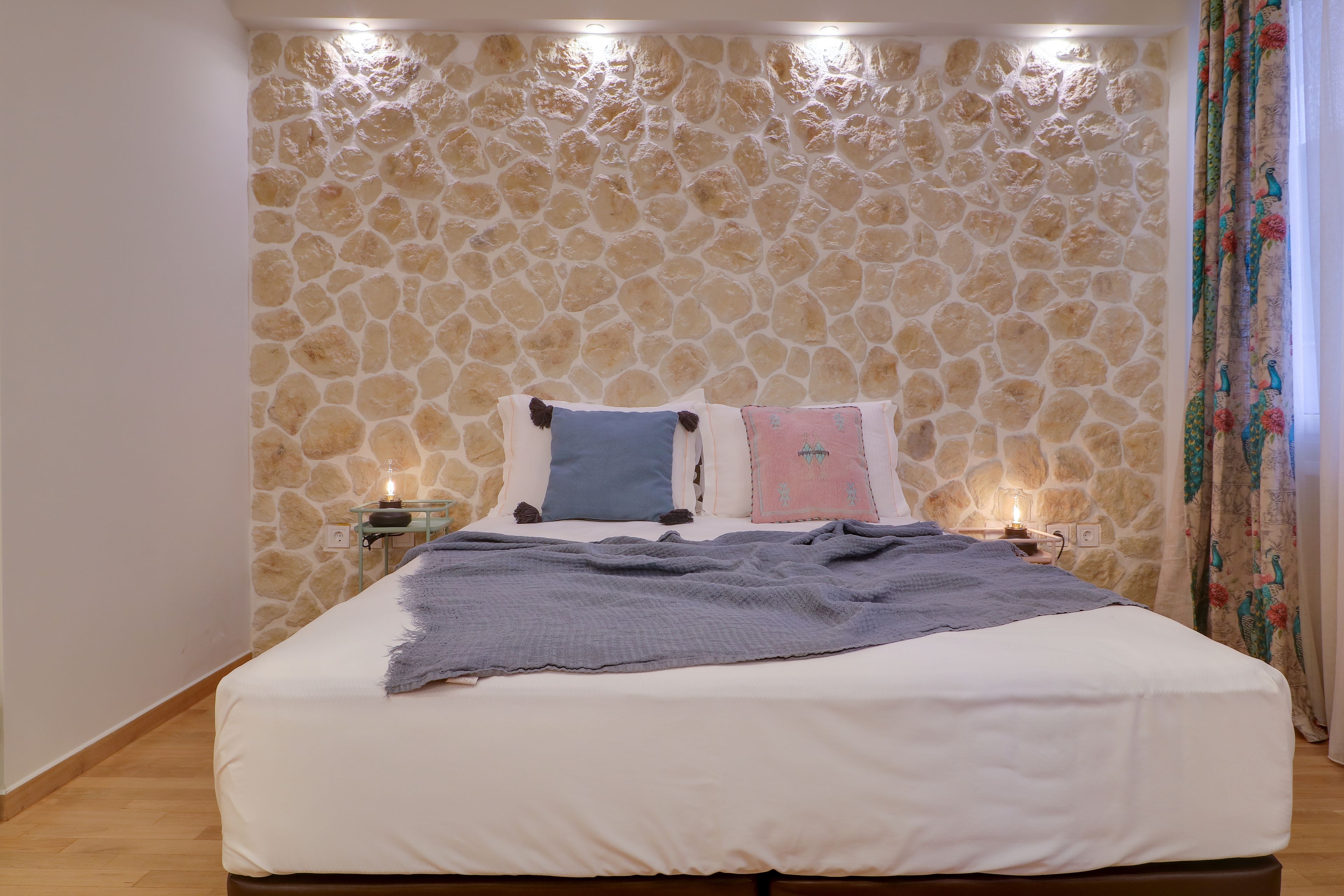 A bedroom with a stone wall, a bed with white bedding, and a nightstand with a lamp.