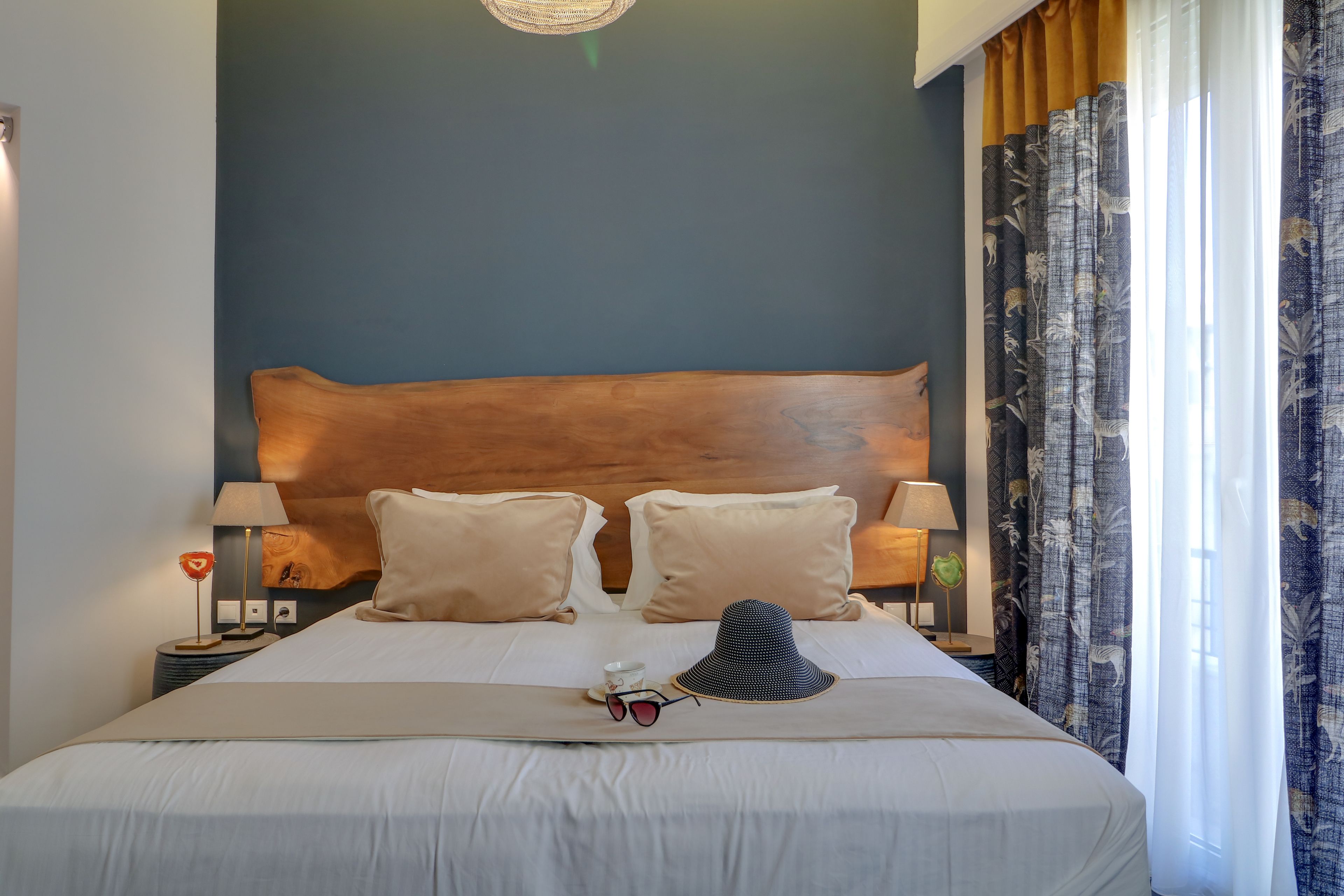A neatly made bed with a wooden headboard, two bedside lamps, and a hat and sunglasses on the bed.