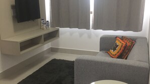 Ein modernes Wohnzimmer mit grauem Sofa, weißem Couchtisch, schwarzem Teppich, Flachbildfernseher und grauem Vorhang.