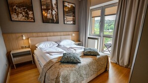 A bedroom with a large bed, a nightstand, a window with a view, and two framed pictures on the wall. - Apartamenty Sun & Snow Pułaskiego (Krynica-Zdroj)