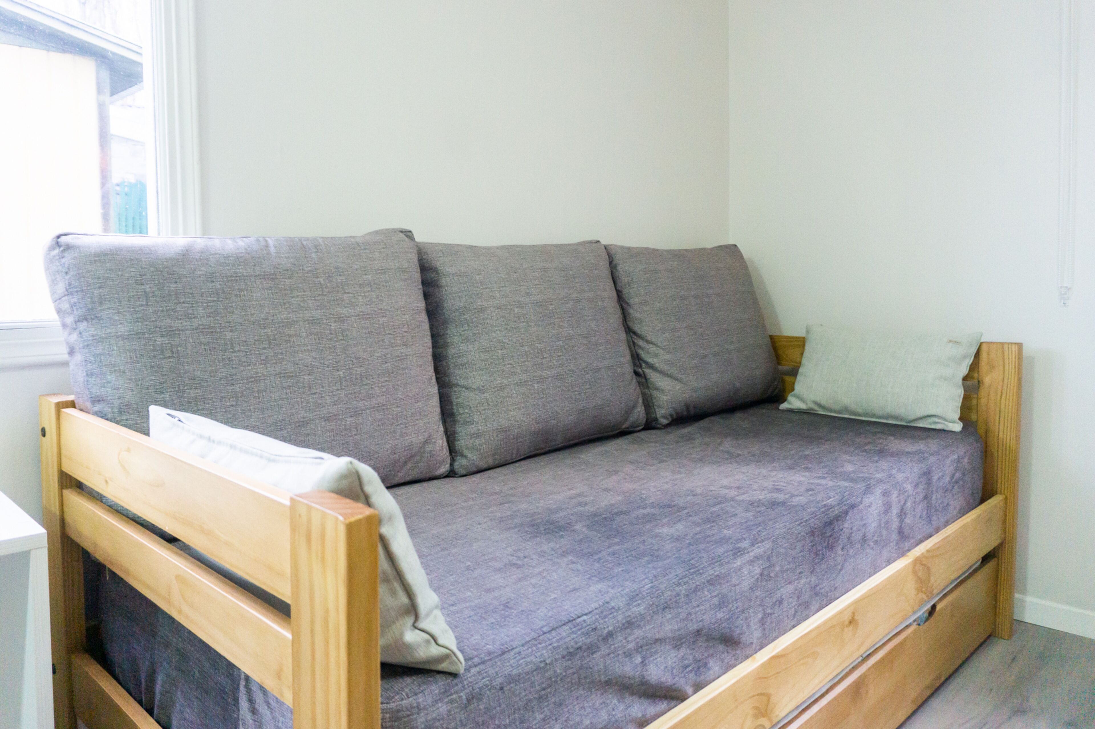 A small, single-bed room with a wooden frame, grey bedding, and a window providing natural light.