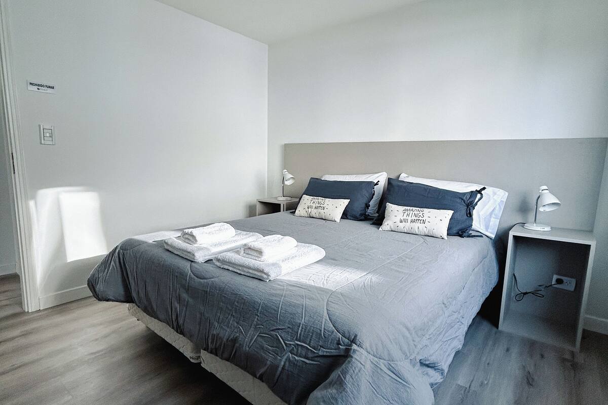 Ein modernes Schlafzimmer mit einem großen Bett, grauer Bettwäsche und einem Nachttisch mit einer Lampe.