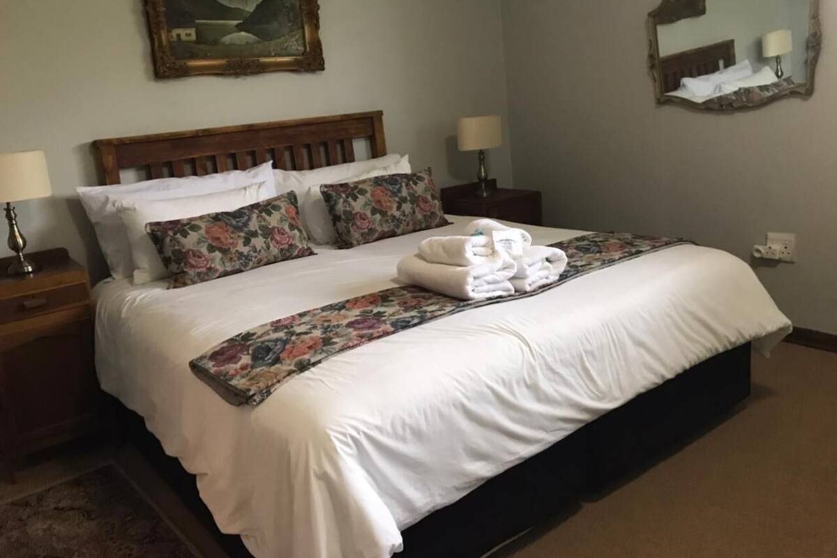 Een net opgemaakt bed met witte lakens, een bloemenbedekking en een stapje opgevouwen handdoeken. Er is een houten hoofdbord, twee nachtkastjes met lampen en een spiegel aan de muur.