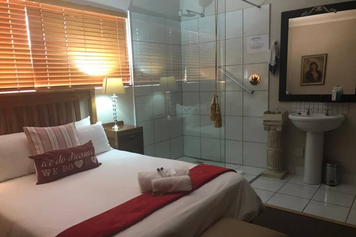 Hotelkamer met een bed, een rode plaid, een kussen met een tekst, een nachtkastje met een lamp, een glazen douchecabine, een badkamer met een wastafel en spiegel.