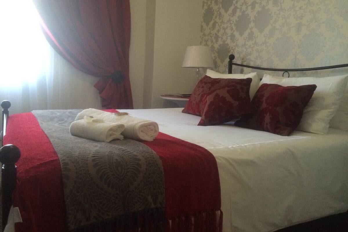 Een net opgemaakt bed met een rood-witte dekbedovertrek, twee rode kussens en een opgerolde witte handdoek aan de zijkant.
