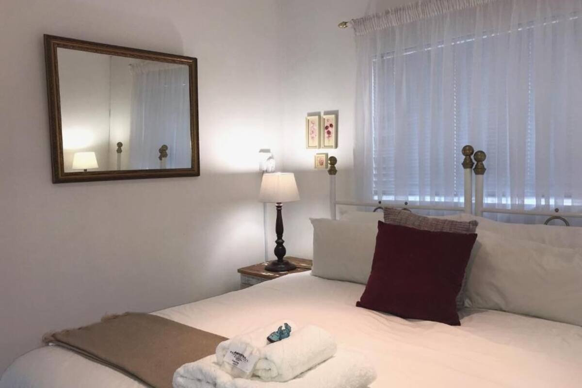 Een hotelkamer met een bed, nachtkastje, lamp, raam met gordijnen en een spiegel aan de muur.