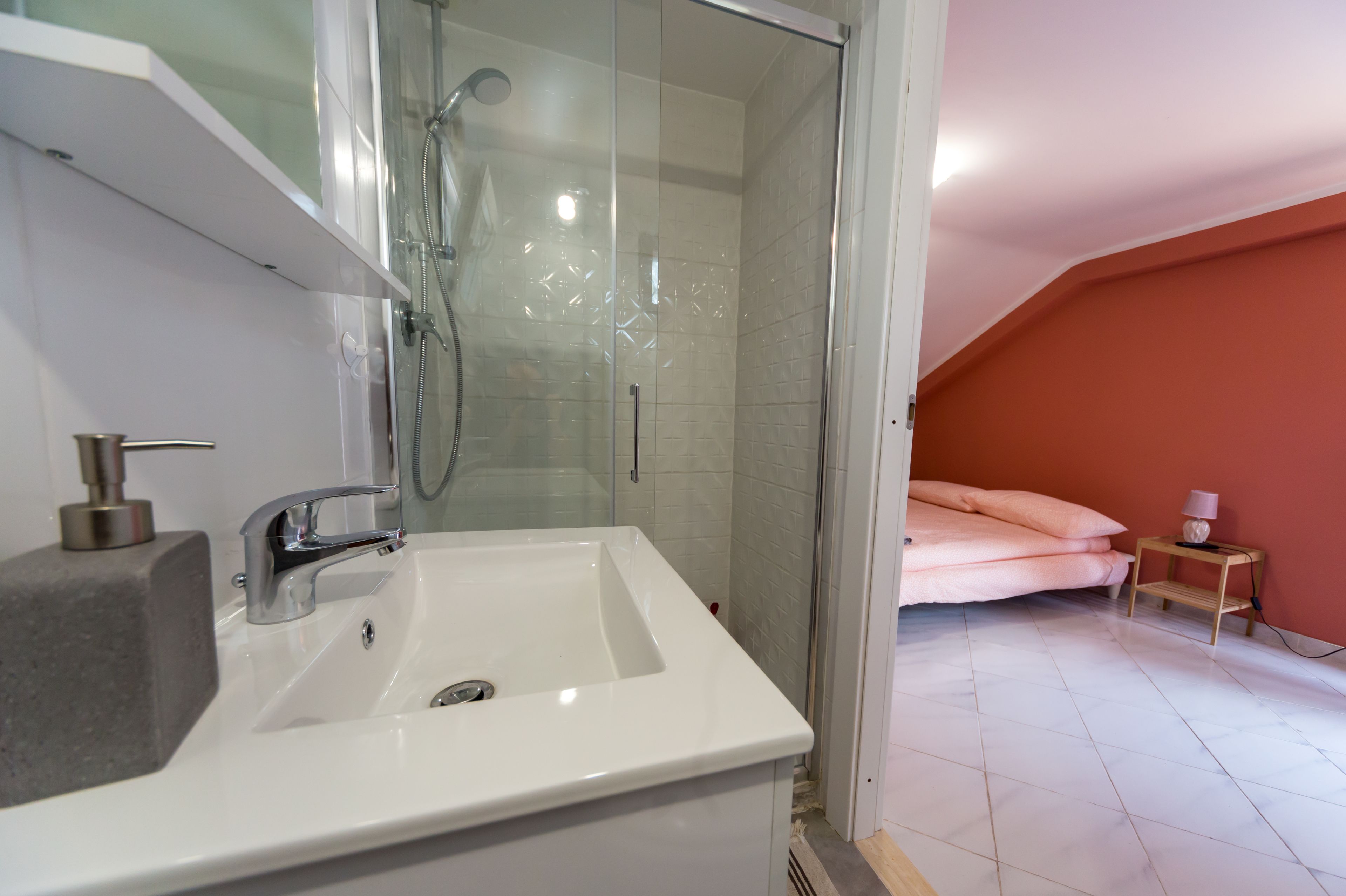 Casa de banho com lavatório branco, cabine de duche com vidro e cama com roupa de cama cor-de-rosa.