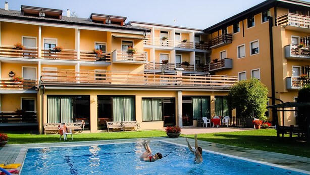 A swimming pool with two people in the water, surrounded by a building with balconies and a lawn area. - Hotel Dolomiti Levico (Levico Terme)