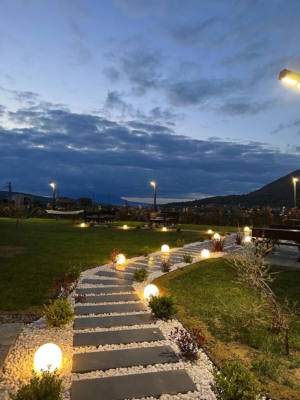 A pathway leading to a seating area with overhead lighting, surrounded by greenery and a view of a distant hill. - Porta Trakis Hotel (Gökçeada)