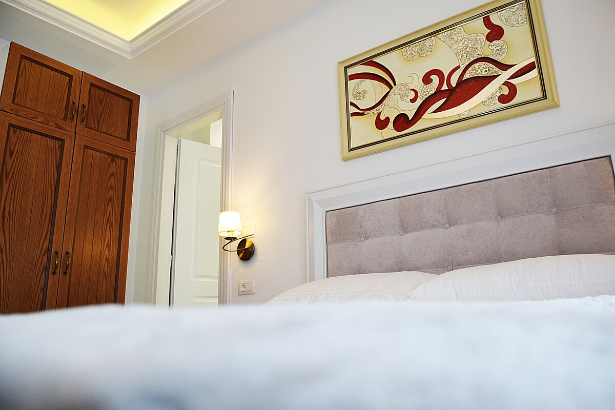 A bedroom with a bed, a wooden wardrobe, a wall-mounted light, and a decorative piece above the bed.