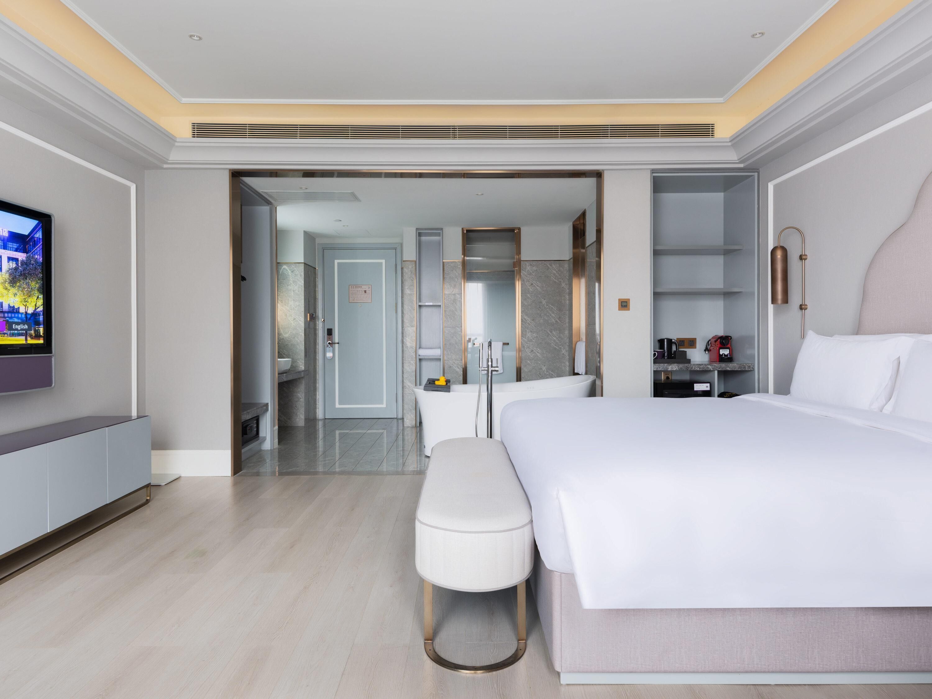 Ein modernes Hotelzimmer mit einem großen Bett, einem Fernseher, einem Schreibtisch und einem durchgegangene Tür einsehbaren Badezimmer.
