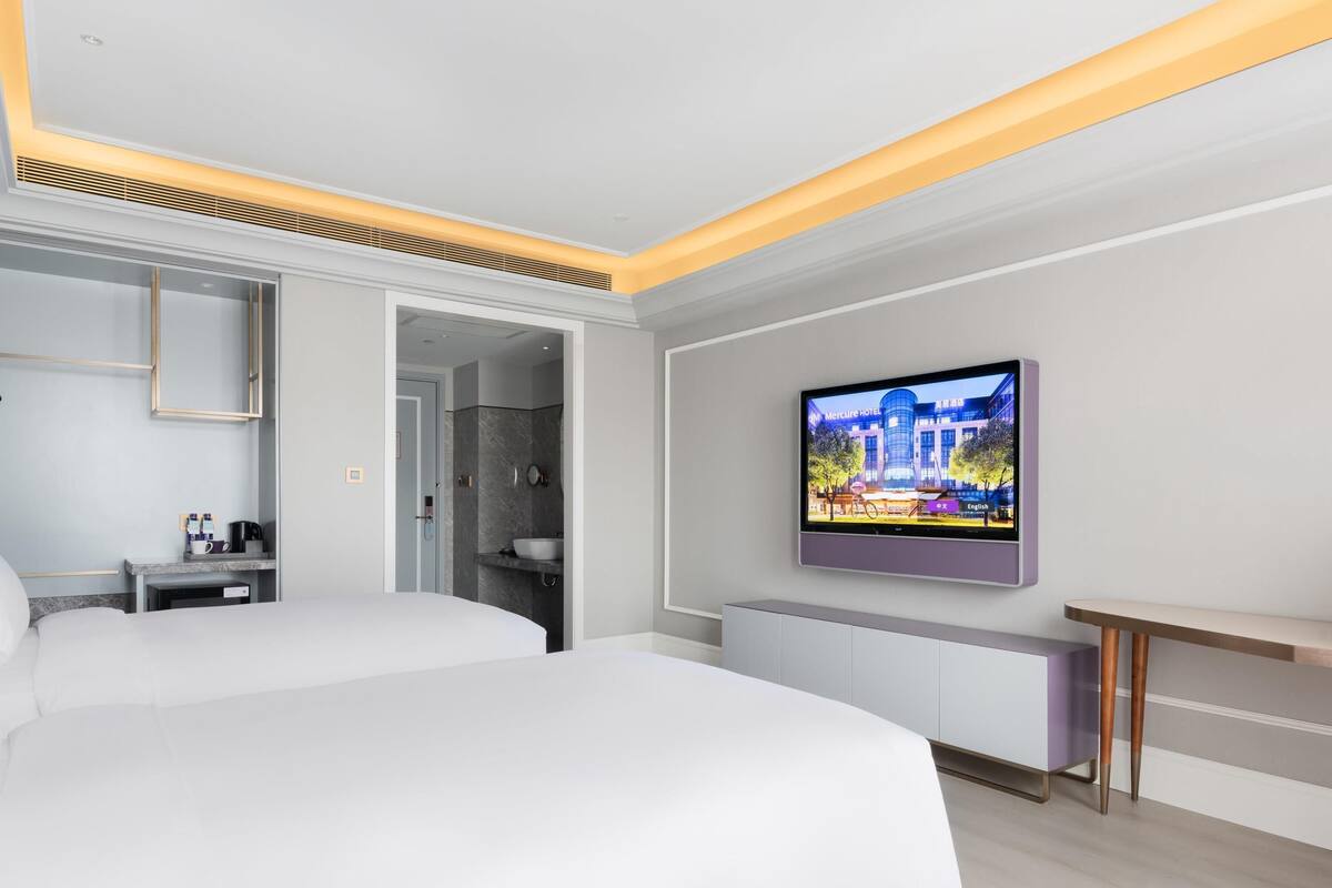 A modern hotel room with a large bed, a TV mounted on the wall, a bedside table with a lamp, and a bathroom visible through an open door.