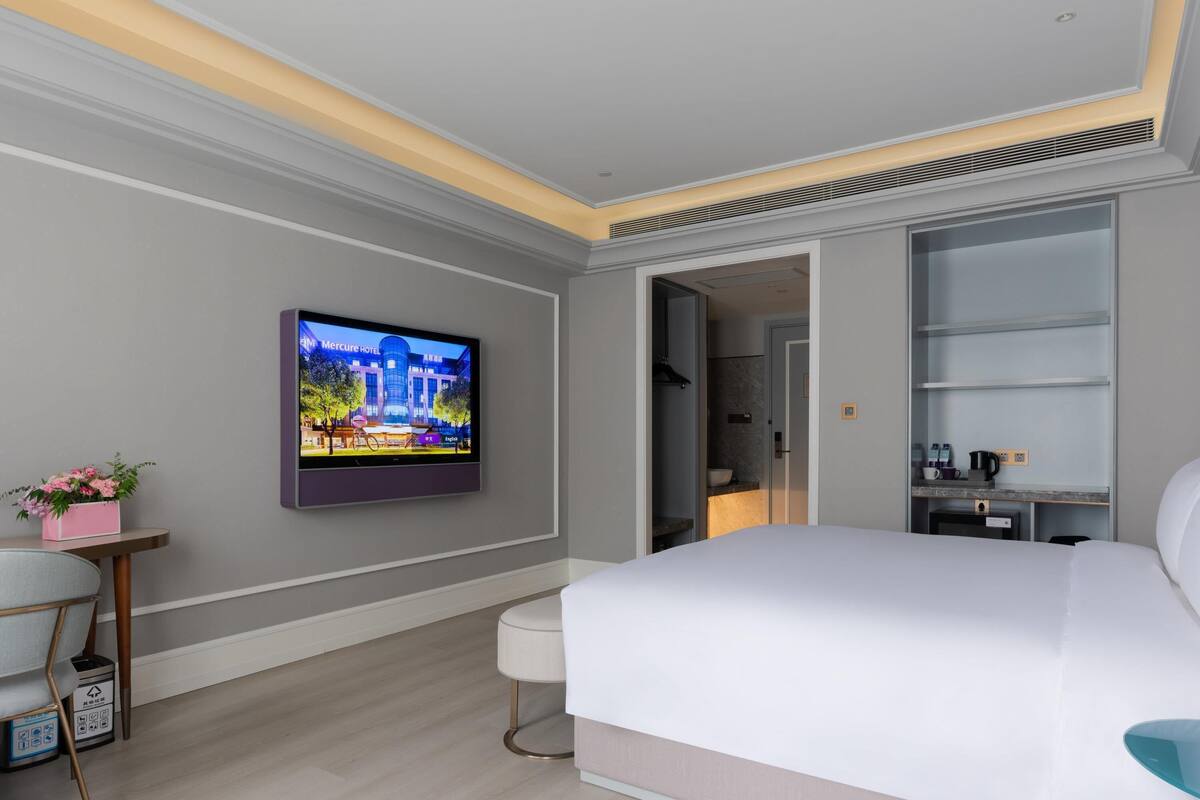 A modern hotel room with a large bed, a television mounted on the wall, a small table with a pink flower vase, and a built-in shelf with toiletries.
