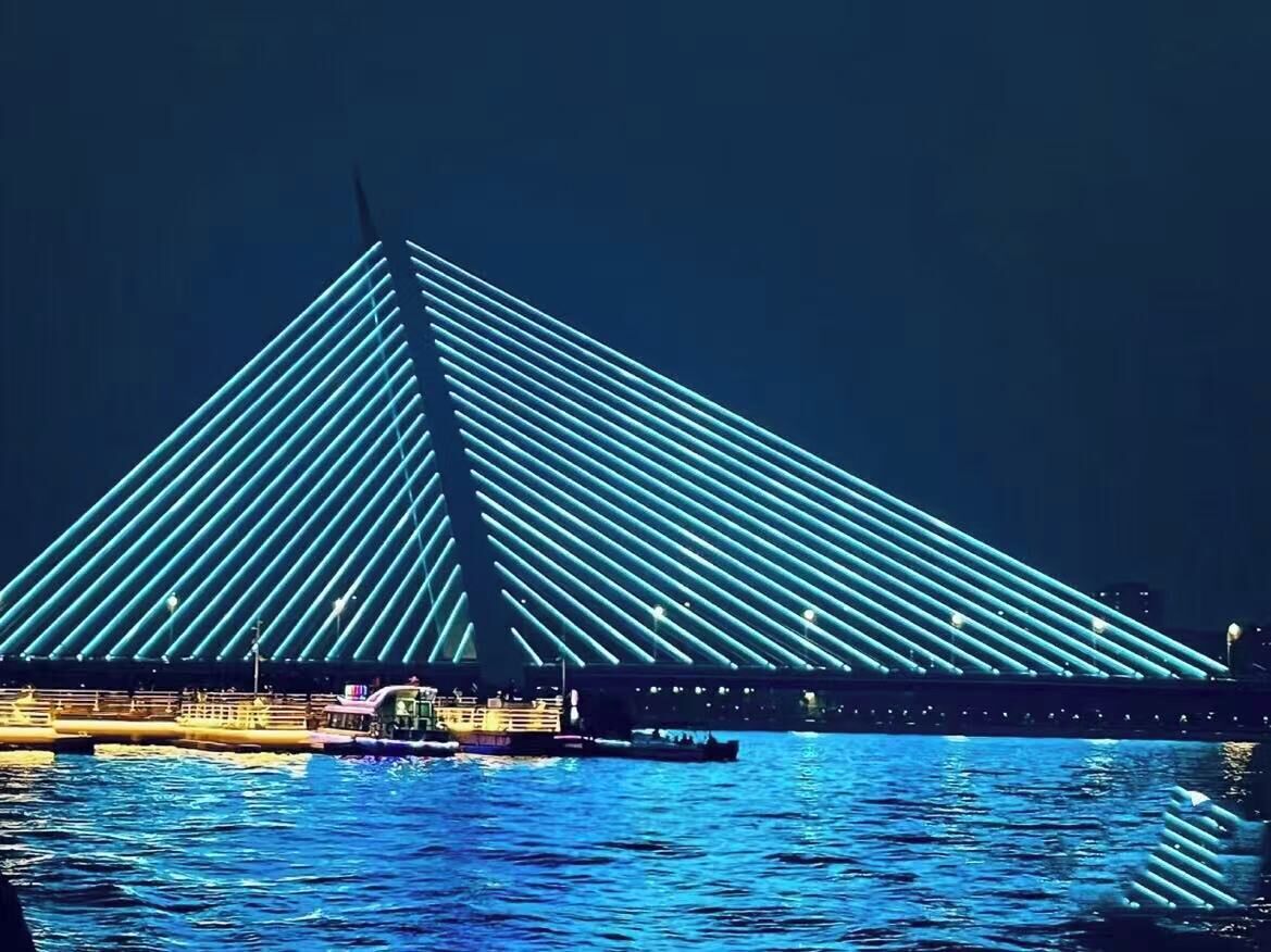 A large, illuminated bridge structure with a triangular design, spanning over a body of water.