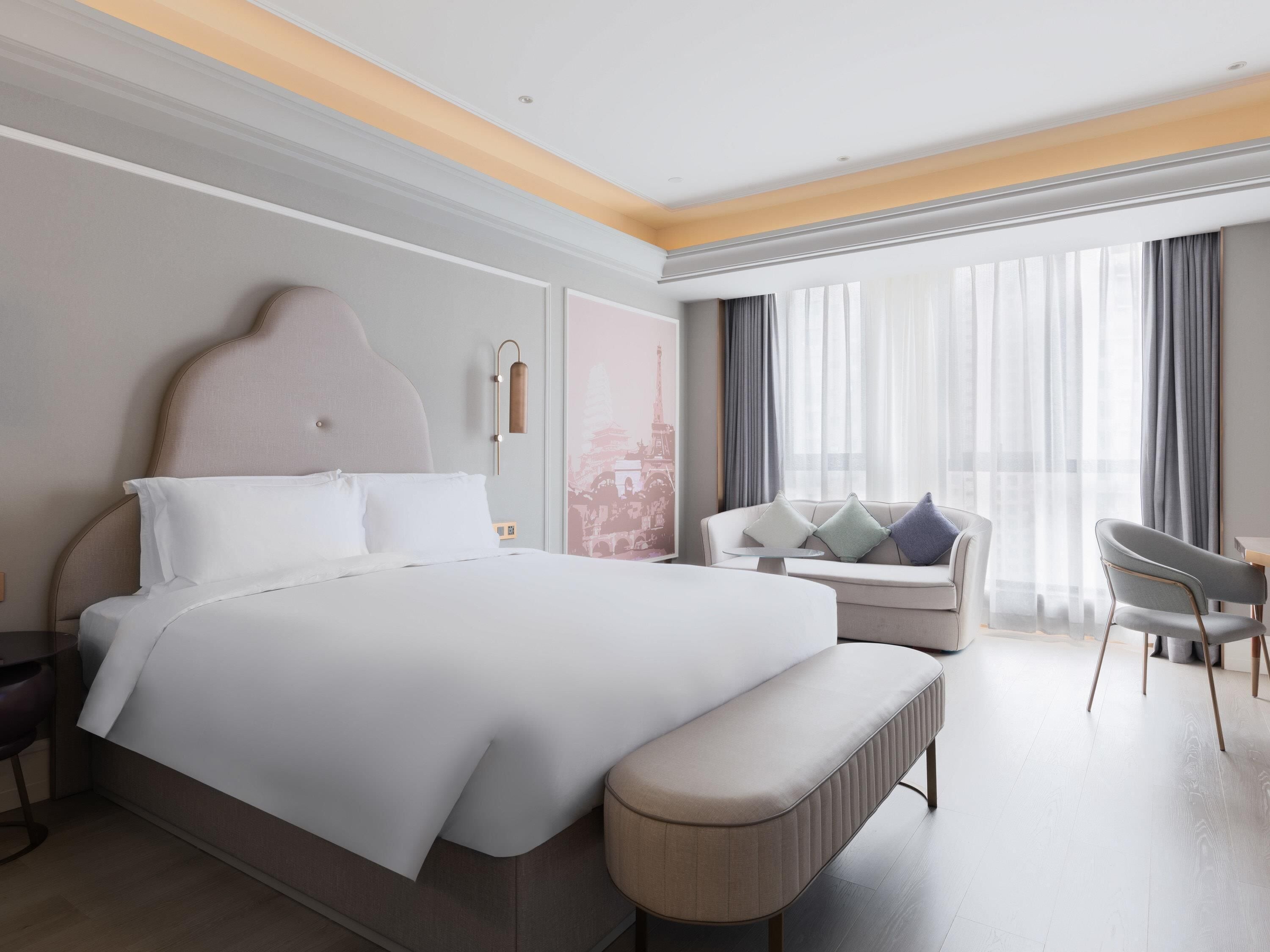 Quarto de hotel moderno com uma cama grande, área de estar com sofá e cadeiras, e uma janela ampla com cortinas.