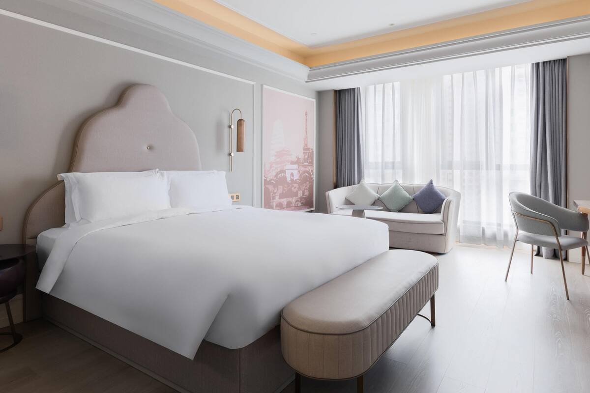 A modern hotel room with a large bed, a seating area with a sofa and chairs, and a large window with curtains.