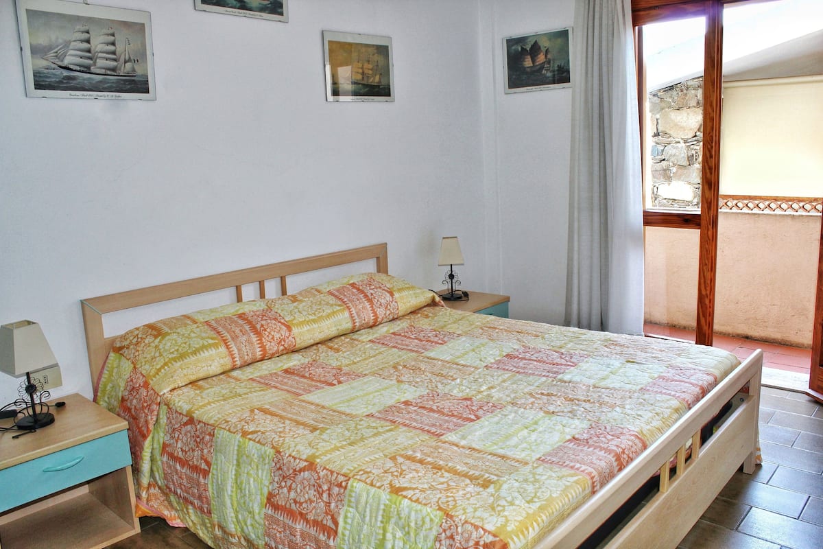 Ein Schlafzimmer mit einem hölzernen Bett, einem Nachttisch, einer Lampe und einem Fenster mit Blick auf eine Steinmauer.