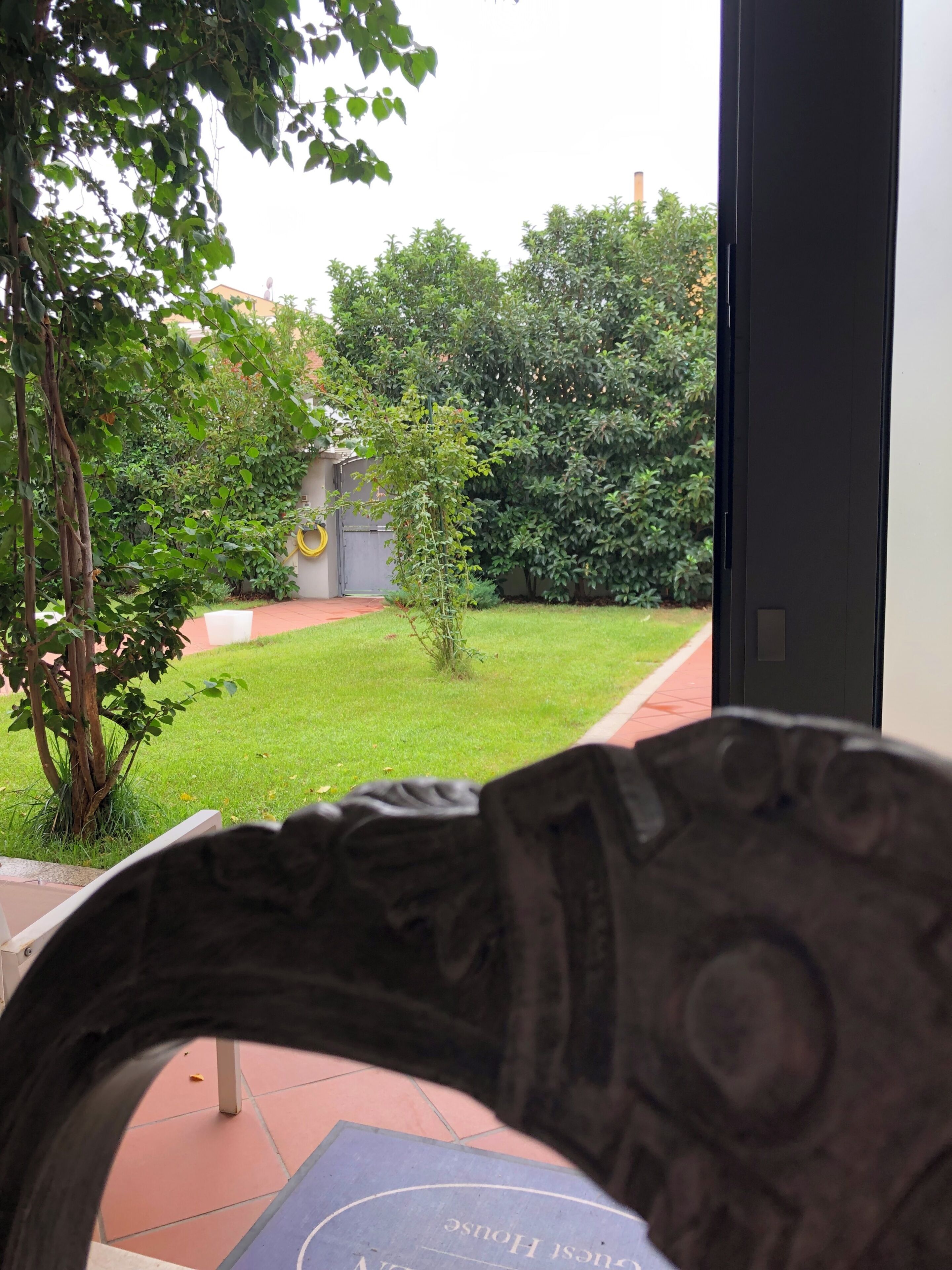 Vista a través de una ventana que muestra un cuarto con una puerta, un pequeño jardín con césped y árboles, y un edificio al fondo.