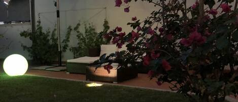 Un patio con un gran paraguas, una mesa redonda y una silla. Se observa un pequeño jardín con un árbol y algunas flores.